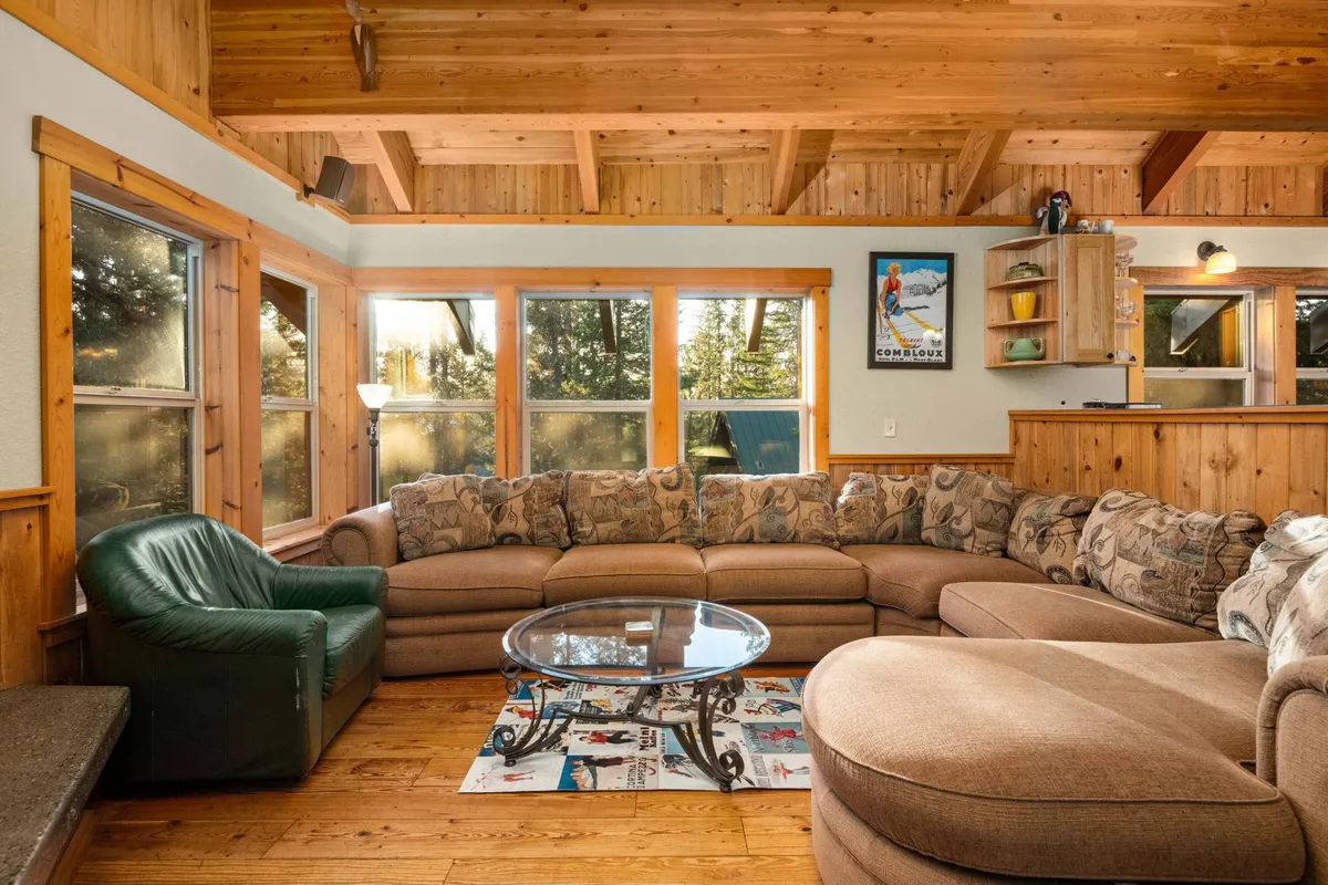 A spacious great room with a large sectional sofa, glass coffee table, wooden ceiling beams, and expansive windows. The space feels inviting and is well-suited for group relaxation, conversation, or casual gatherings.