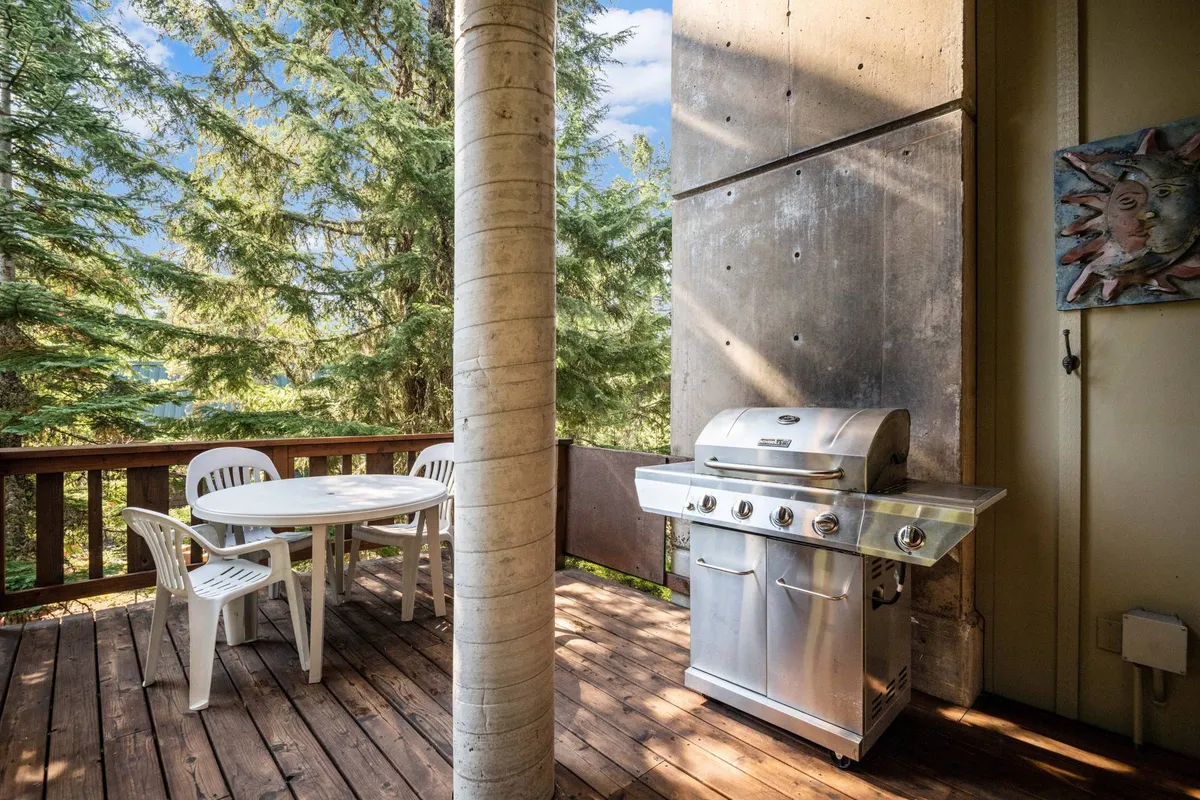 An inviting wooden deck set amid tall trees, featuring a gas grill and an outdoor dining table and chairs. The scene suggests a relaxed atmosphere ideal for summer barbecues and outdoor meals after a day of adventure.