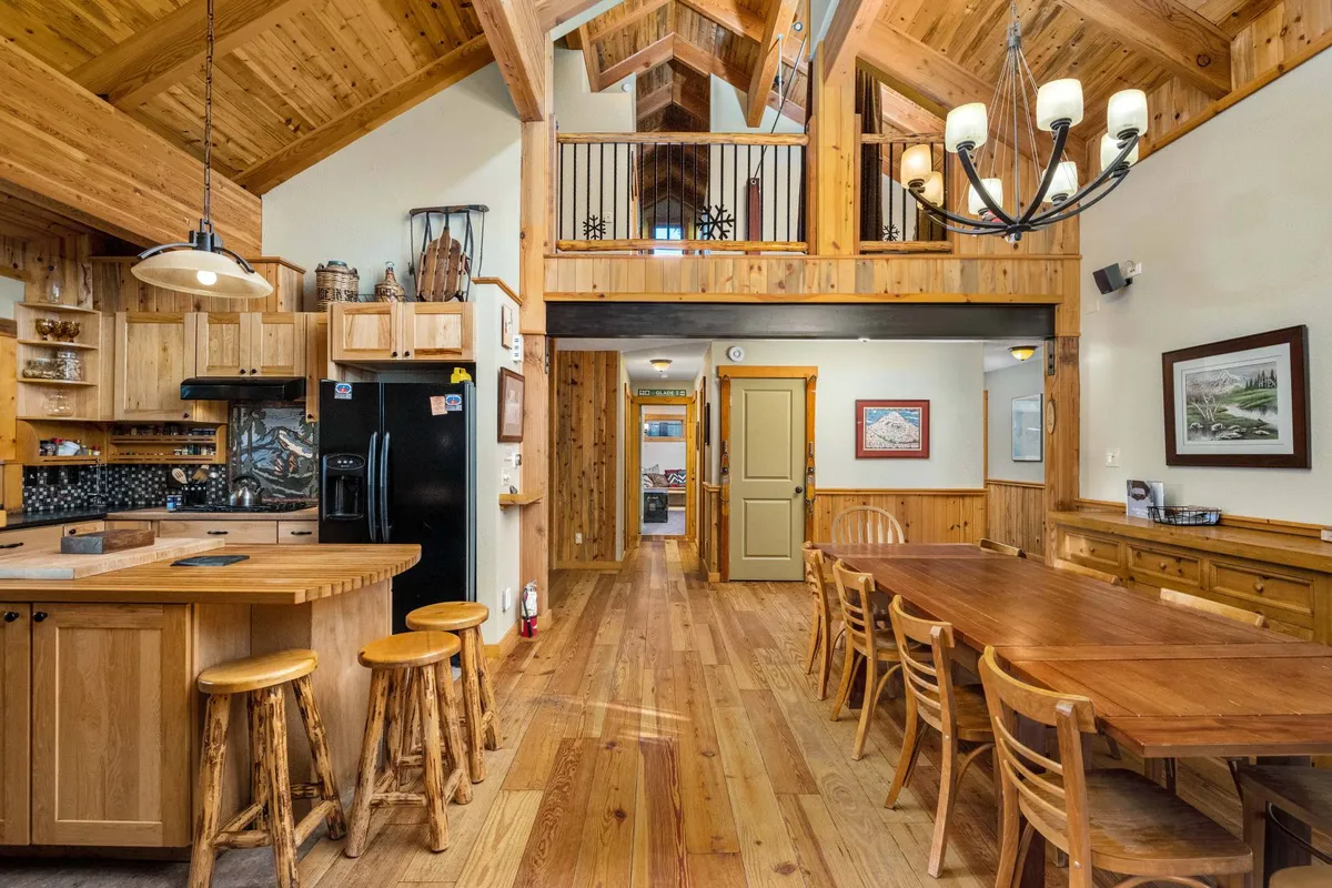 A wide shot of the dining area and kitchen, featuring a large wooden dining table beneath a high vaulted ceiling, a kitchen island, chandelier, and an open loft area above. The space is bathed in warm light, inviting communal meals and lively gatherings.
