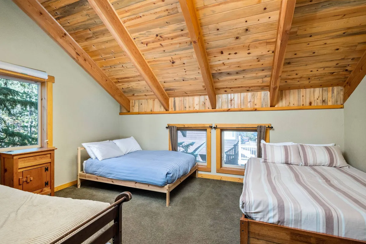 A spacious bedroom with exposed wooden beams, large windows letting in natural sunlight, and inviting bedding arrangements. The setting exudes a warm, comfortable atmosphere perfect for relaxation, reading, or playful evenings. Timber elements in the architecture and the snow outside the windows reinforce the luxe cabin ambiance.