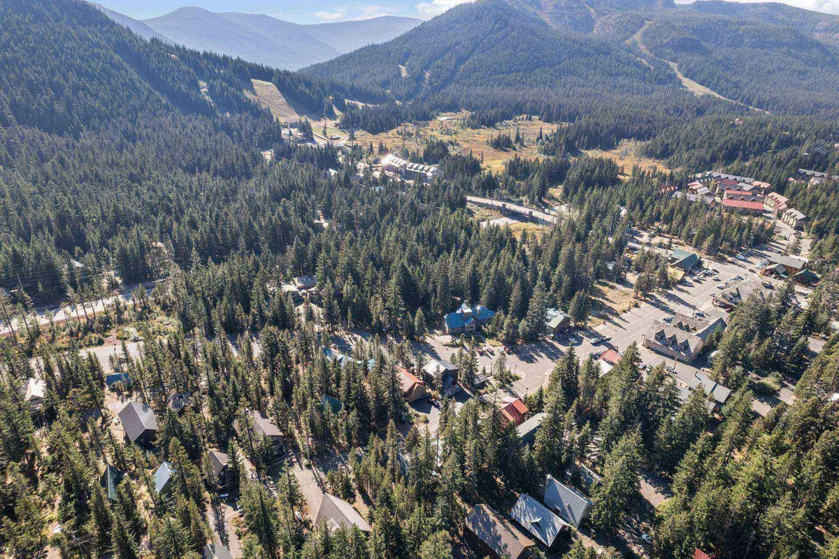 A panoramic view of a dense forest, sprawling open spaces, scattered cabins, and a dramatic mountain range in the distance. The lighting suggests early evening or sunset, heightening the sense of tranquility and natural wonder.