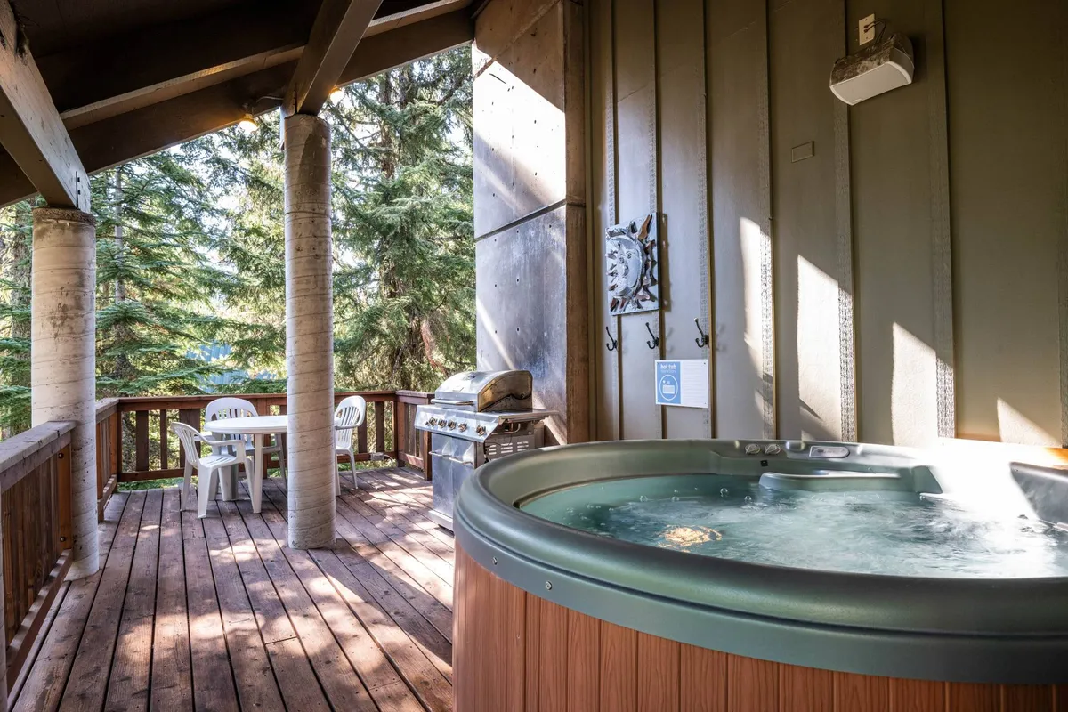 A serene exterior deck scene featuring a bubbling hot tub, a gas grill, a wooden deck, and an outdoor dining table surrounded by chairs. Lush trees envelop the space, and soft sunset lighting hints at the beauty of evenings spent outdoors.