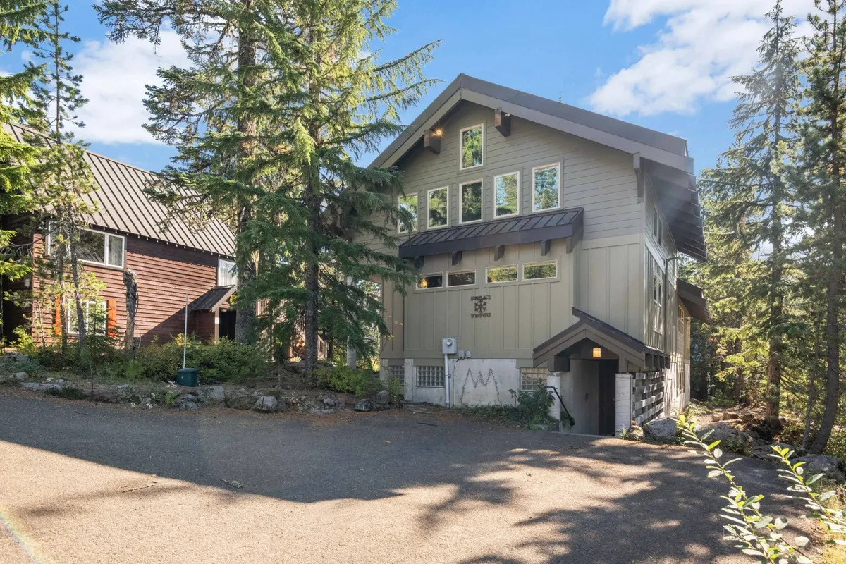 The image focuses on the main entrance of the property, featuring expansive windows and mature trees enveloping the building. The sun-drenched porch suggests a seamless transition between indoor comfort and outdoor village exploration.