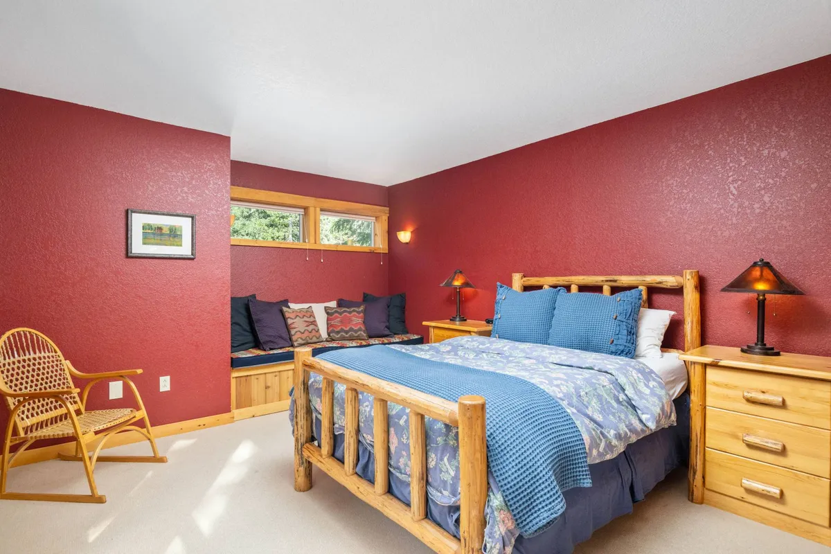 A bedroom featuring a wooden bed with blue bedding, a window seat filled with cushions, and matching wooden nightstands with warm lamps. Natural light pours in through the windows, spotlighting the tranquil, inviting setting.