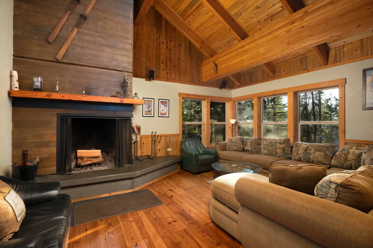 The image features a spacious, inviting living area with a roaring fireplace, large picture windows showcasing forested views, and plush, comfortable seating arranged for groups or families. The room glows with warm lighting, creating a cozy atmosphere perfect for gathering together after a day on the slopes.