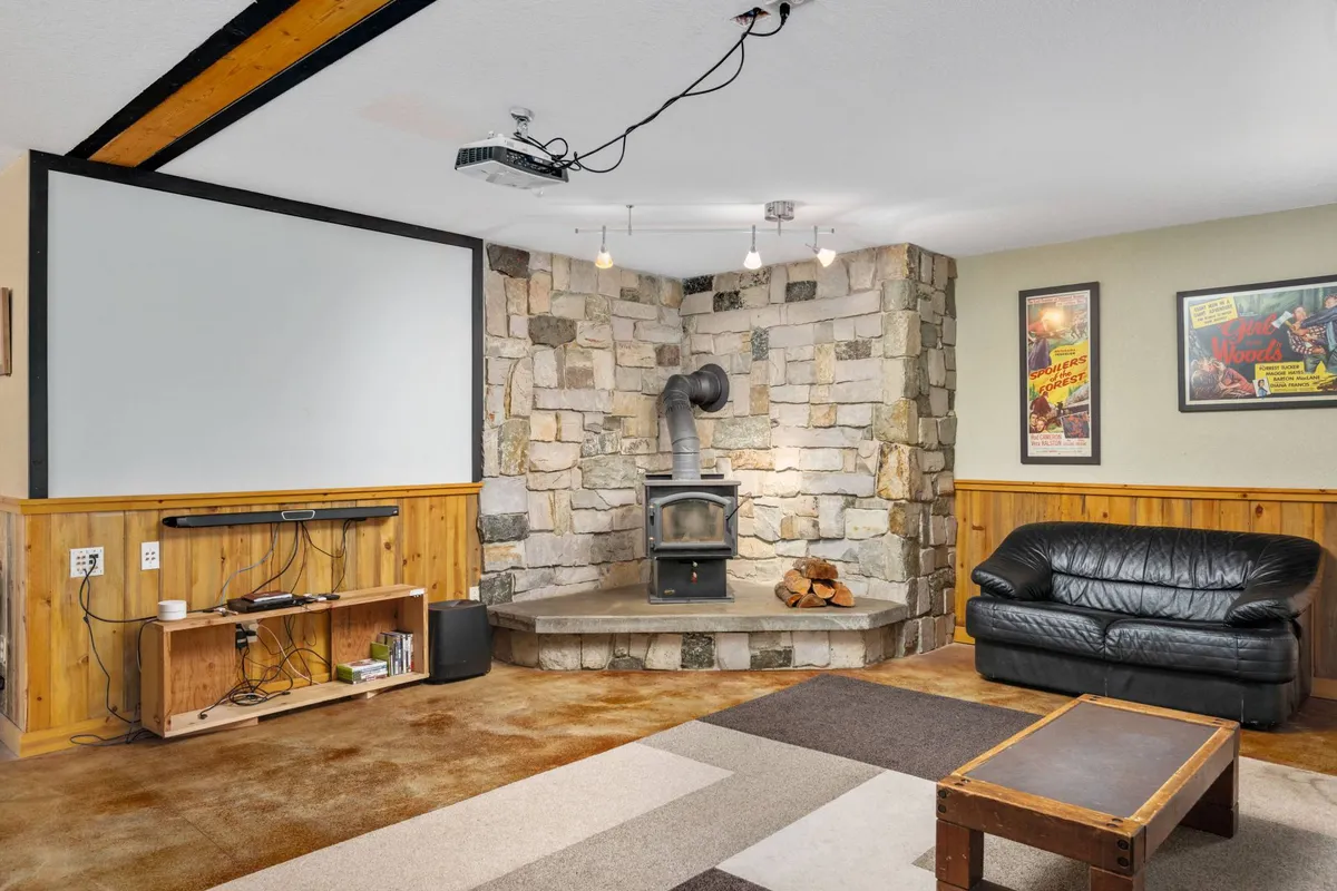 A cozy home theater area featuring a stone fireplace, projector screen, black leather couch, and a wooden coffee table. The setting is perfect for movie nights, games, or relaxed evenings together.