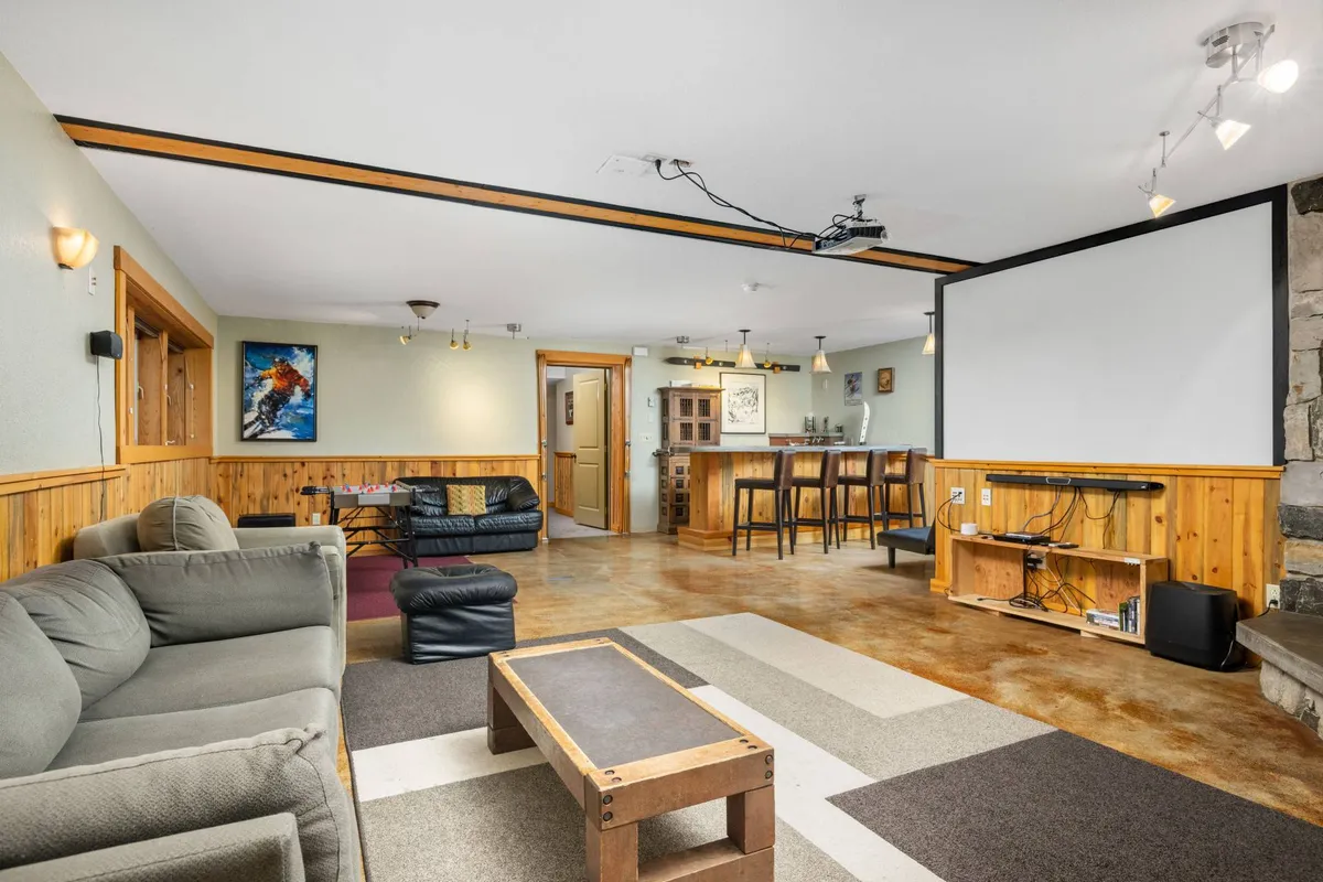 A spacious, comfortable entertainment room featuring a projector screen for movie nights, plush seating for groups, a sleek bar counter, and vibrant artwork on the wall. The layout supports both cinematic experiences and casual gatherings.